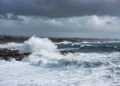 Onde potenti e spruzzi bianchi si infrangono sulla costa rocciosa di Marina di Ragusa sotto un cielo grigio e ventoso, previsione meteo Pasqua 2026 con maltempo sugli Iblei