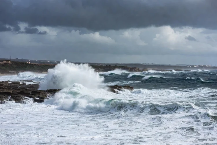 Onde potenti e spruzzi bianchi si infrangono sulla costa rocciosa di Marina di Ragusa sotto un cielo grigio e ventoso, previsione meteo Pasqua 2026 con maltempo sugli Iblei