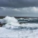 Onde potenti e spruzzi bianchi si infrangono sulla costa rocciosa di Marina di Ragusa sotto un cielo grigio e ventoso, previsione meteo Pasqua 2026 con maltempo sugli Iblei