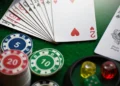 Una vista ravvicinata di un ventaglio di carte da gioco, chip da poker impilate con denominazioni (5, 10, 20), e dadi colorati (rosso, giallo, verde) su un tavolo da gioco in feltro verde.