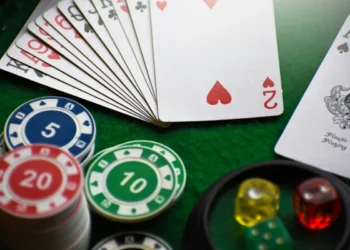 Una vista ravvicinata di un ventaglio di carte da gioco, chip da poker impilate con denominazioni (5, 10, 20), e dadi colorati (rosso, giallo, verde) su un tavolo da gioco in feltro verde.