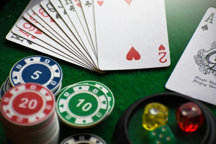 Una vista ravvicinata di un ventaglio di carte da gioco, chip da poker impilate con denominazioni (5, 10, 20), e dadi colorati (rosso, giallo, verde) su un tavolo da gioco in feltro verde.