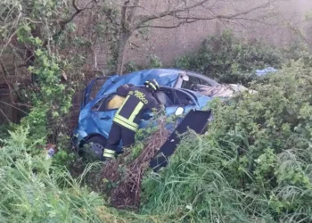 Un vigile del fuoco con uniforme "Vigili del Fuoco" è chino su una Citroen C3 blu gravemente danneggiata e accartocciata, finita nella fitta vegetazione di una scarpata in contrada Monachella, Ragusa.