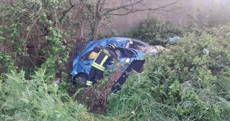 Un vigile del fuoco con uniforme "Vigili del Fuoco" è chino su una Citroen C3 blu gravemente danneggiata e accartocciata, finita nella fitta vegetazione di una scarpata in contrada Monachella, Ragusa.
