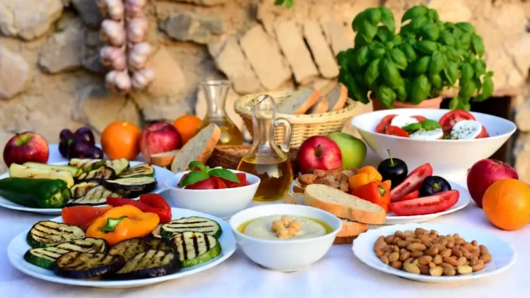 Una tavola imbandita con piatti tipici della dieta mediterranea: verdure grigliate (zucchine, melanzane, peperoni), hummus di ceci, insalata caprese, pane integrale, frutta fresca, mandorle e olio d'oliva su uno sfondo rustico in pietra.