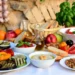 Una tavola imbandita con piatti tipici della dieta mediterranea: verdure grigliate (zucchine, melanzane, peperoni), hummus di ceci, insalata caprese, pane integrale, frutta fresca, mandorle e olio d'oliva su uno sfondo rustico in pietra.
