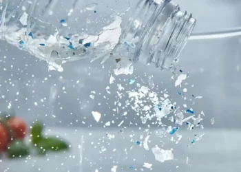Primo piano macro di una bottiglia di plastica che si sbriciola rilasciando microparticelle e frammenti azzurri in una caraffa d'acqua limpida, con verdure fresche sfocate sullo sfondo.
