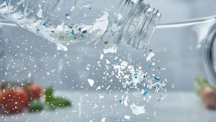 Primo piano macro di una bottiglia di plastica che si sbriciola rilasciando microparticelle e frammenti azzurri in una caraffa d'acqua limpida, con verdure fresche sfocate sullo sfondo.