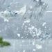 Primo piano macro di una bottiglia di plastica che si sbriciola rilasciando microparticelle e frammenti azzurri in una caraffa d'acqua limpida, con verdure fresche sfocate sullo sfondo.