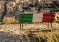 Una panchina dipinta con il tricolore italiano si trova in una zona panoramica di Modica Alta, con la città sullo sfondo. Un piccolo logo di Fratelli d'Italia e il testo dell'evento sono visibili.