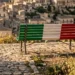 Una panchina dipinta con il tricolore italiano si trova in una zona panoramica di Modica Alta, con la città sullo sfondo. Un piccolo logo di Fratelli d'Italia e il testo dell'evento sono visibili.