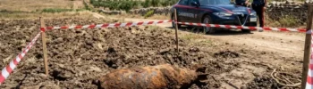 Un vecchio ordigno bellico arrugginito adagiato su un terreno agricolo appena arato, delimitato da nastro segnaletico bianco e rosso. Sullo sfondo, una pattuglia dei Carabinieri e due militari in servizio in una zona rurale.