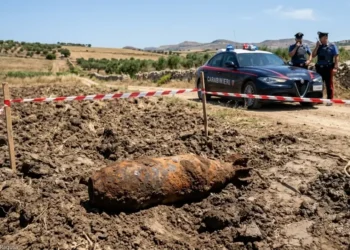 Un vecchio ordigno bellico arrugginito adagiato su un terreno agricolo appena arato, delimitato da nastro segnaletico bianco e rosso. Sullo sfondo, una pattuglia dei Carabinieri e due militari in servizio in una zona rurale.