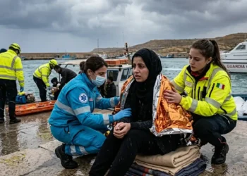Una giovane donna migrante con uno sguardo traumatizzato, avvolta in una coperta termica dorata, assistita da soccorritori al molo Favarolo di Lampedusa. Sullo sfondo, una motovedetta della Guardia Costiera e operatori sanitari in un contesto di maltempo.