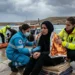 Una giovane donna migrante con uno sguardo traumatizzato, avvolta in una coperta termica dorata, assistita da soccorritori al molo Favarolo di Lampedusa. Sullo sfondo, una motovedetta della Guardia Costiera e operatori sanitari in un contesto di maltempo.