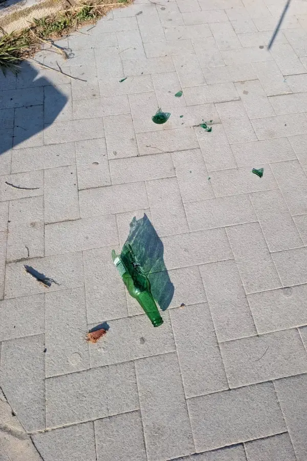 Una bottiglia di birra di vetro verde rotta e diversi frammenti di vetro sparsi su un marciapiede lastricato grigio in una giornata di sole.