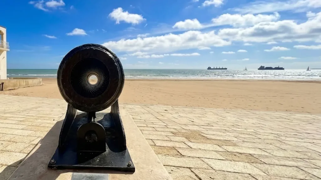Primo piano della bocca di un antico cannone in ghisa sul lungomare di Marina di Ragusa, puntato verso la spiaggia dorata e il Mar Mediterraneo sotto un cielo azzurro