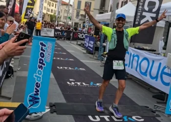 Michele Sammito esulta a braccia aperte e dita in segno di vittoria mentre taglia il traguardo della UTLAC 250k in Piazza XX Settembre a Lecco. L'atleta indossa una divisa tecnica, pettorale numero 53 e zaino da trail, circondato dal pubblico e dai banner dell'evento.