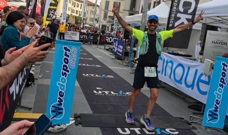 Michele Sammito esulta a braccia aperte e dita in segno di vittoria mentre taglia il traguardo della UTLAC 250k in Piazza XX Settembre a Lecco. L'atleta indossa una divisa tecnica, pettorale numero 53 e zaino da trail, circondato dal pubblico e dai banner dell'evento.