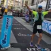 Michele Sammito esulta a braccia aperte e dita in segno di vittoria mentre taglia il traguardo della UTLAC 250k in Piazza XX Settembre a Lecco. L'atleta indossa una divisa tecnica, pettorale numero 53 e zaino da trail, circondato dal pubblico e dai banner dell'evento.