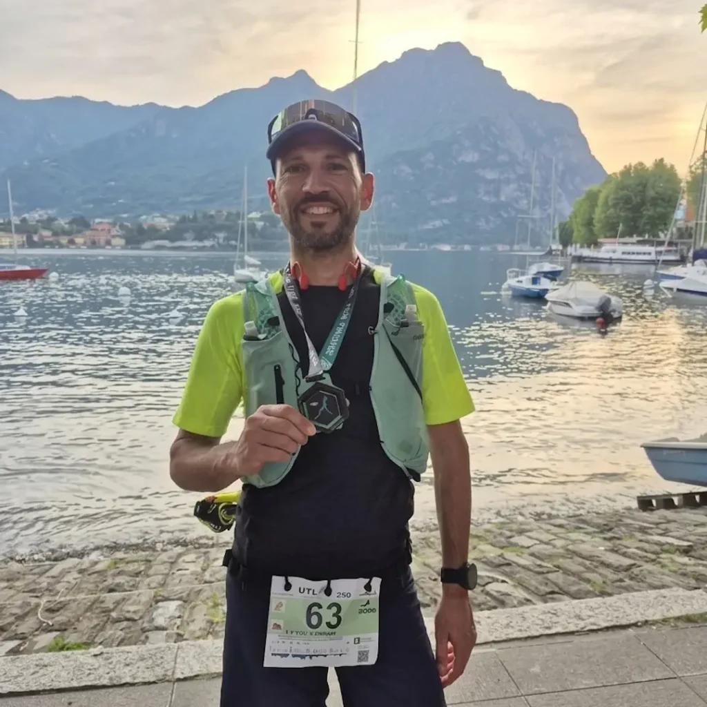 Un primo piano di Michele Sammito sorridente mentre mostra la medaglia di finisher della UTLAC 250k. Indossa abbigliamento tecnico, zaino da trail e il pettorale numero 63. Sullo sfondo il Lago di Como al tramonto con montagne e barche.