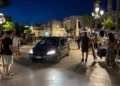 Auto all'interno di un'area pedonale affollata durante le ore serali, con cittadini che osservano la scena in una piazza di Marina di Ragusa