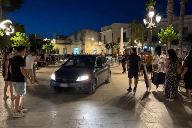 Auto all'interno di un'area pedonale affollata durante le ore serali, con cittadini che osservano la scena in una piazza di Marina di Ragusa