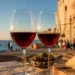 Immagine editoriale generata con intelligenza artificiale: calici di vino rosso Cerasuolo su tavolo con prodotti tipici siciliani, sullo sfondo la Torre Cabrera e il mare di Pozzallo durante la rassegna Calici dei Cabrera