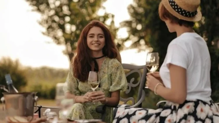 Due donne con abiti estivi sorseggiano un aperitivo all'aperto durante una serata estiva, con look leggeri e informali