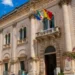 Facciata del Palazzo Municipale di Scicli, sede del Comune, con le bandiere italiana, europea e siciliana all'ingresso.