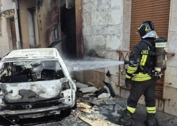Un Vigile del Fuoco interviene per spegnere l'auto completamente distrutta dalle fiamme in via Rattazzi a Vittoria, dove il rogo ha danneggiato anche il portone di un'abitazione