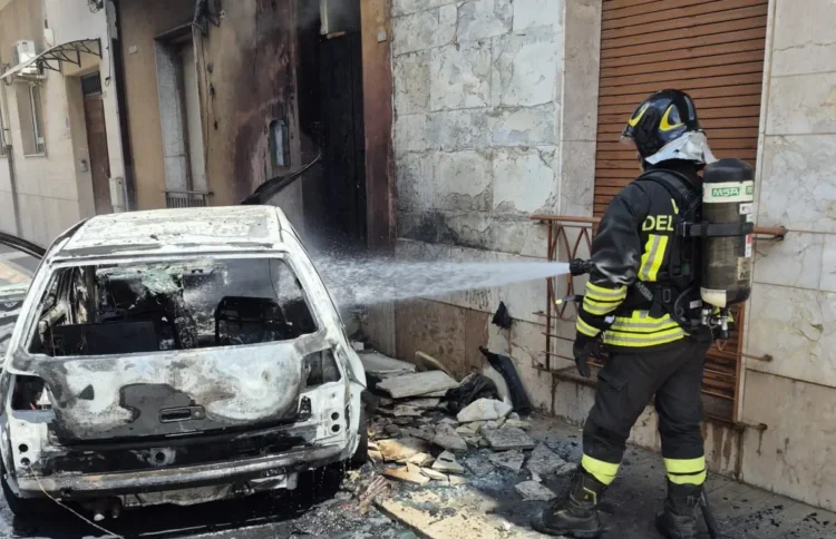Un Vigile del Fuoco interviene per spegnere l'auto completamente distrutta dalle fiamme in via Rattazzi a Vittoria, dove il rogo ha danneggiato anche il portone di un'abitazione