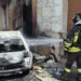 Un Vigile del Fuoco interviene per spegnere l'auto completamente distrutta dalle fiamme in via Rattazzi a Vittoria, dove il rogo ha danneggiato anche il portone di un'abitazione