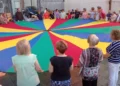 Partecipanti riuniti attorno a un grande paracadute colorato durante un momento di animazione in piazza Carmine, nell'ambito della festa della Madonna del Carmine a Ragusa.