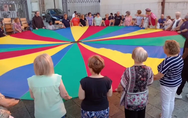 Partecipanti riuniti attorno a un grande paracadute colorato durante un momento di animazione in piazza Carmine, nell'ambito della festa della Madonna del Carmine a Ragusa.