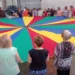 Partecipanti riuniti attorno a un grande paracadute colorato durante un momento di animazione in piazza Carmine, nell'ambito della festa della Madonna del Carmine a Ragusa.