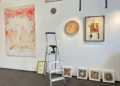 Allestimento della mostra "Mostri e altri Maestri" alla Galleria Quam di Scicli con opere d'arte contemporanea, una scala utilizzata durante la preparazione e disegni di bambini esposti lungo la parete.