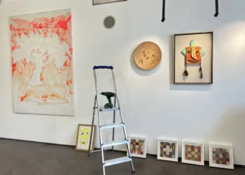 Allestimento della mostra "Mostri e altri Maestri" alla Galleria Quam di Scicli con opere d'arte contemporanea, una scala utilizzata durante la preparazione e disegni di bambini esposti lungo la parete.