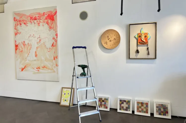 Allestimento della mostra "Mostri e altri Maestri" alla Galleria Quam di Scicli con opere d'arte contemporanea, una scala utilizzata durante la preparazione e disegni di bambini esposti lungo la parete.