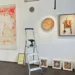 Allestimento della mostra "Mostri e altri Maestri" alla Galleria Quam di Scicli con opere d'arte contemporanea, una scala utilizzata durante la preparazione e disegni di bambini esposti lungo la parete.