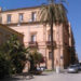 Palazzo storico di Vittoria con facciata color ocra, palme e strada laterale con auto parcheggiate