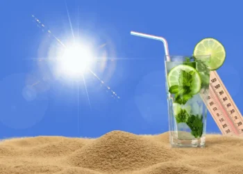 Bicchiere con lime e menta su sabbia, accanto a un termometro che segna oltre 40°C, cielo azzurro con sole intenso