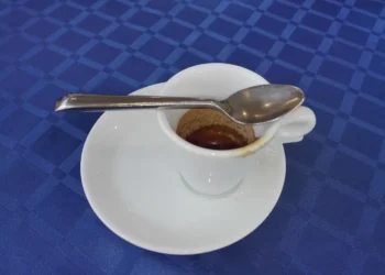Tazzina di caffè espresso quasi vuota con cucchiaino sul piattino bianco, su tovaglia a quadri blu.