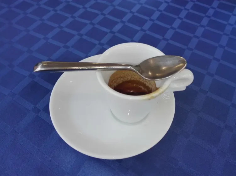 Tazzina di caffè espresso quasi vuota con cucchiaino sul piattino bianco, su tovaglia a quadri blu.