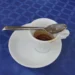 Tazzina di caffè espresso quasi vuota con cucchiaino sul piattino bianco, su tovaglia a quadri blu.