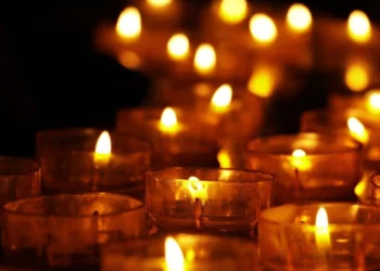 Primo piano di diverse candele votive accese, con fiamme che illuminano un ambiente buio, simbolo di lutto, commemorazione e raccoglimento.