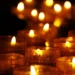 Primo piano di diverse candele votive accese, con fiamme che illuminano un ambiente buio, simbolo di lutto, commemorazione e raccoglimento.