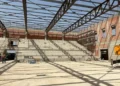 Interno del nuovo palazzetto dello sport di Comiso in costruzione, con gradinate in cemento, struttura portante della copertura e operai presenti durante il sopralluogo dell'assessore ai Lavori pubblici.