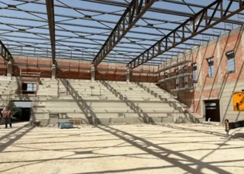 Interno del nuovo palazzetto dello sport di Comiso in costruzione, con gradinate in cemento, struttura portante della copertura e operai presenti durante il sopralluogo dell'assessore ai Lavori pubblici.
