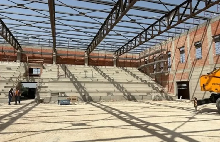 Interno del nuovo palazzetto dello sport di Comiso in costruzione, con gradinate in cemento, struttura portante della copertura e operai presenti durante il sopralluogo dell'assessore ai Lavori pubblici.